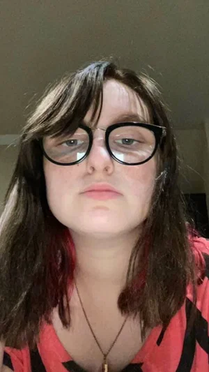 Woman with dark hair and glasses wearing a red and black top indoors.