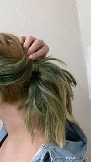 Person with green-tinted hair tying a messy bun, wearing denim jacket indoors.