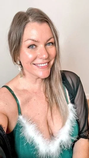 Woman with blonde hair in green feathered top and sheer black sleeves, smiling indoors.