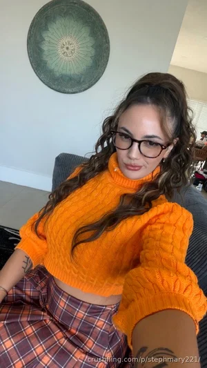 Woman with curly hair and glasses wears orange sweater and plaid skirt, sitting indoors.