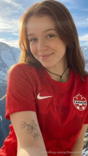 Woman in red Canada soccer jersey with tattoo on arm, smiling outdoors with snowy mountains behind.