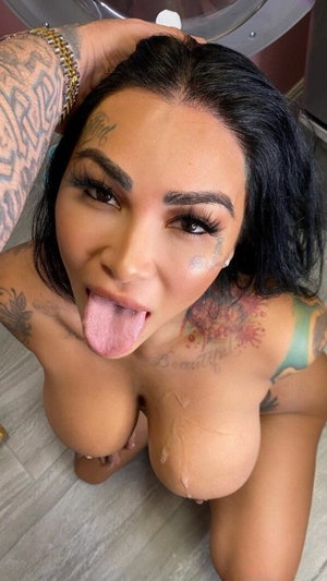 Woman with tattoos and dark hair posing topless indoors, tongue out, hand on head.