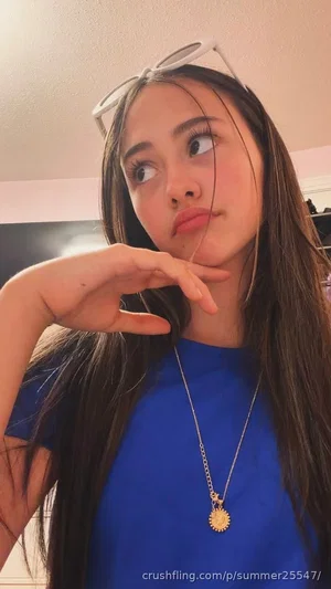 Woman with long dark hair wearing blue t-shirt and gold pendant, posing indoors with sunglasses on