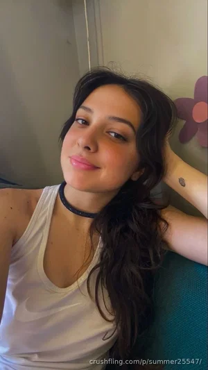 Woman with long dark hair in white tank top, smiling, indoors.
