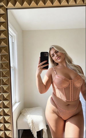 Woman with blonde hair in pink lingerie taking mirror selfie in bedroom.