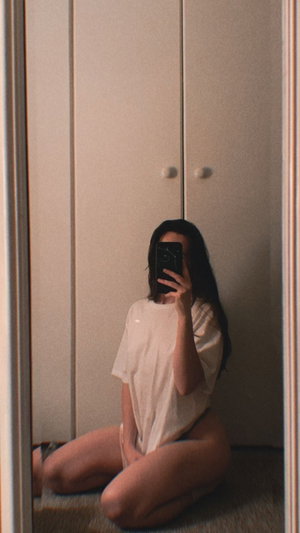 Woman with long dark hair sits on floor in loose white shirt, taking mirror selfie in bedroom.
