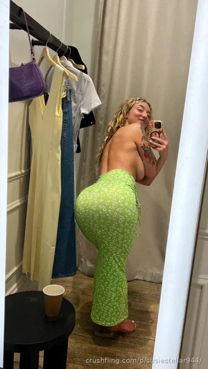 Woman with curly blonde hair in green patterned pants, bare back, taking mirror selfie in bedroom.