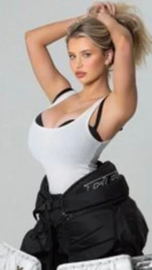 Woman in white tank top and black pants posing with hands in hair, studio setting.