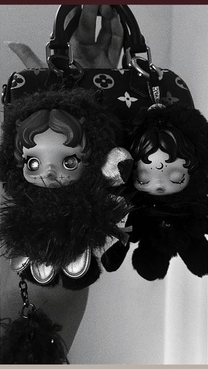 Hand holding a patterned handbag with two doll keychains, black and white photo.