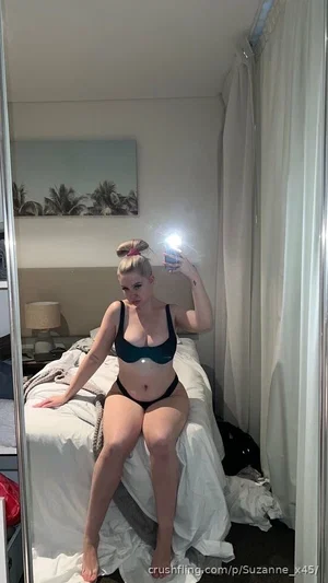 Woman in black lingerie sits on bed, holding phone for mirror selfie in bedroom.
