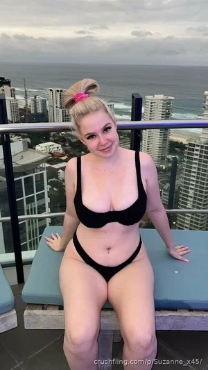 Woman in black bikini sits on lounge chair on balcony overlooking city and ocean.