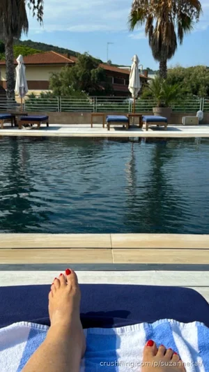 Person's feet with red toenails on towel by poolside, outdoor setting with palm trees and lounge