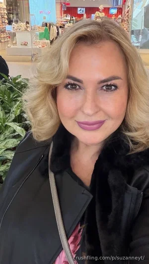 Blonde woman in black jacket with fur collar, smiling in a store.