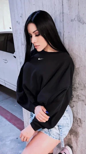 Woman with long black hair in black sweatshirt and denim shorts leaning against concrete wall