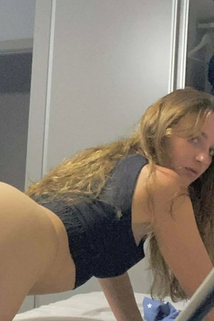 Blonde woman in dark blue outfit posing on bed indoors.