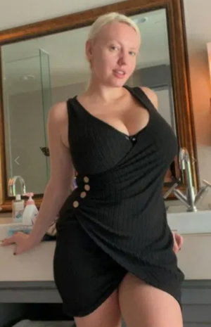 Blonde woman in black dress posing in bathroom.