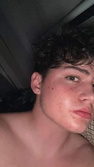 Shirtless man with curly dark hair, close-up selfie in dim indoor setting.
