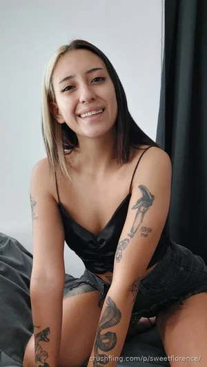 Woman with tattoos in black top and shorts, sitting indoors.