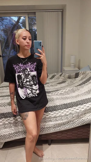 Woman with blonde hair and tattoos wears black graphic tee, stands barefoot in bedroom.