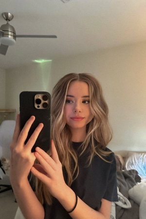 Slim blonde woman in black top takes bedroom selfie.
