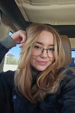 Blonde woman with glasses in denim jacket taking car selfie.