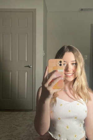 Woman with blonde hair in white top with yellow flowers, taking mirror selfie indoors.