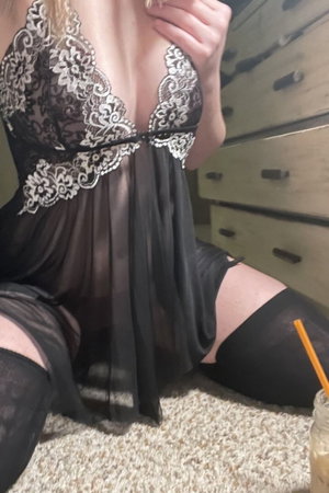Woman in black lace lingerie and thigh-high stockings kneeling on carpet near dresser.