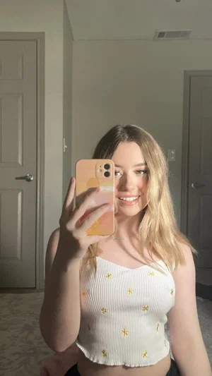Woman with blonde hair in white crop top taking mirror selfie in bedroom.