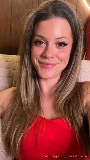 Woman with long brown hair wearing red top, sitting indoors on a beige couch.
