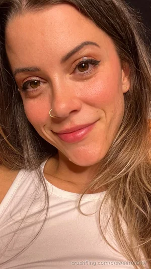Woman with long brown hair and nose ring, wearing white tank top, smiling in close-up selfie.