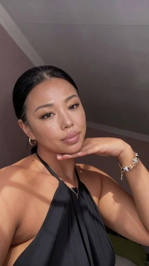 Woman with dark hair in black halter top posing indoors, hand under chin.
