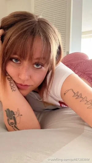 Woman with tattoos on arms, wearing white t-shirt, lying on bed indoors.
