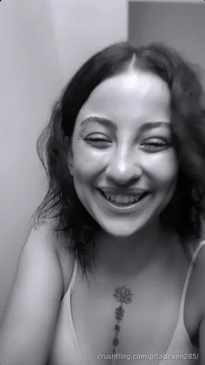 Woman with curly hair, smiling, wearing a tank top, chest tattoo visible, indoor setting.