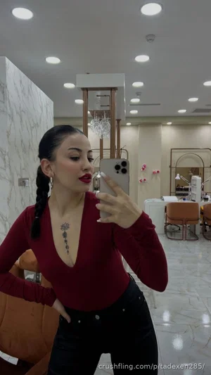 Woman in red top and black pants takes mirror selfie in salon.