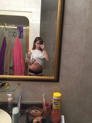 Woman in black underwear and white shirt tied around torso, taking mirror selfie in bathroom.