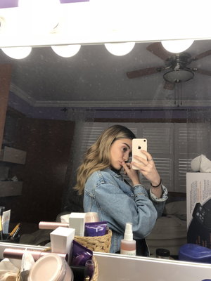 Woman with wavy hair in denim jacket taking mirror selfie in bedroom with makeup items visible.