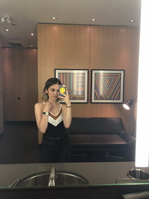 Woman in black and white top and dark pants takes mirror selfie in modern bathroom.