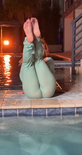 Woman in green leggings sits by pool edge, legs raised, feet together, at night.