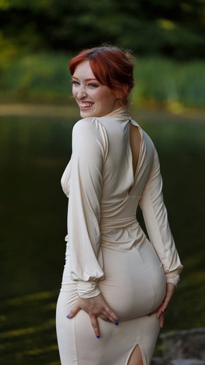 Curvy redhead in white dress by lake outdoors.