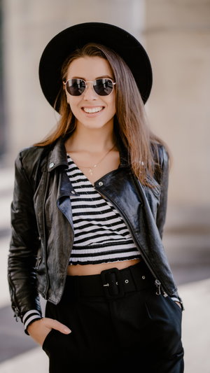 Slim woman in hat, sunglasses, leather jacket, striped crop top, pants outdoors.