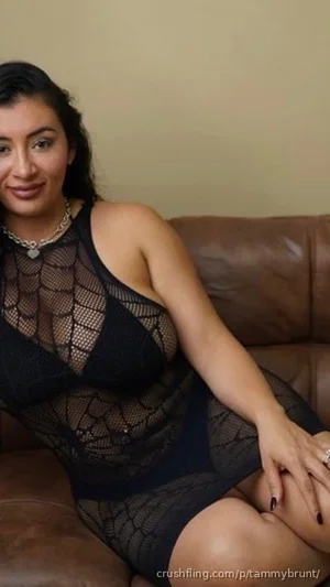 Woman in black mesh bodysuit sitting on brown leather couch, smiling at camera.