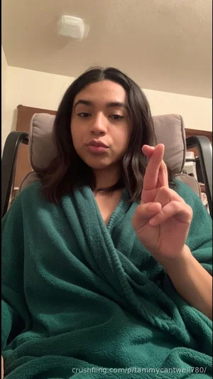 Woman with dark hair in green robe sitting in chair, making hand gesture.