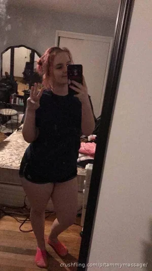Woman with pink hair in black t-shirt and shorts takes mirror selfie in bedroom.