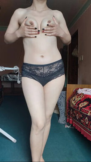 Woman covering breasts with hands wearing lace panties in bedroom.