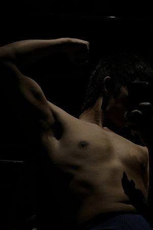 Asian man flexing arm shirtless in underwear dark indoor selfie.