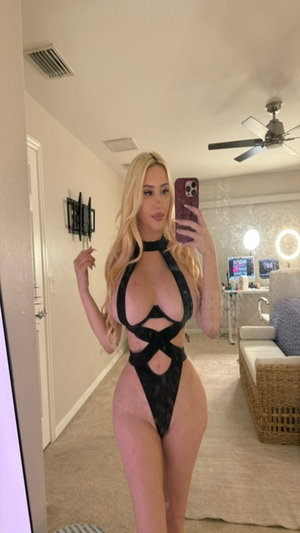 Curvy blonde in black lace bodysuit taking mirror selfie in bedroom.