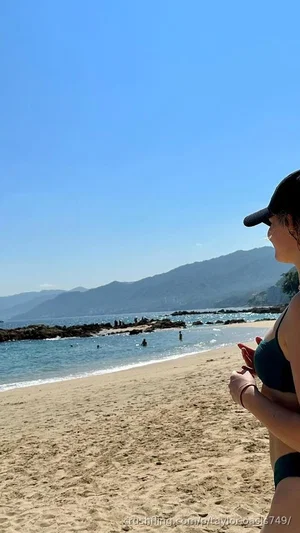 Woman in green bikini standing on sandy beach with ocean and mountains in background.