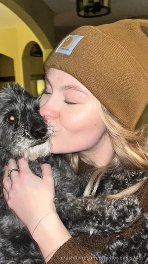 Woman with blonde hair wearing brown beanie and sweater, hugging a curly-haired dog indoors.