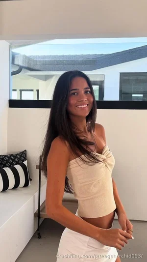 Woman with long dark hair in a beige top and white skirt, smiling indoors near a window.