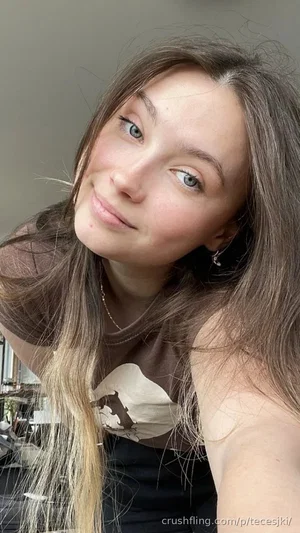 Woman with long brown hair wearing a brown graphic top, smiling in a casual indoor setting.